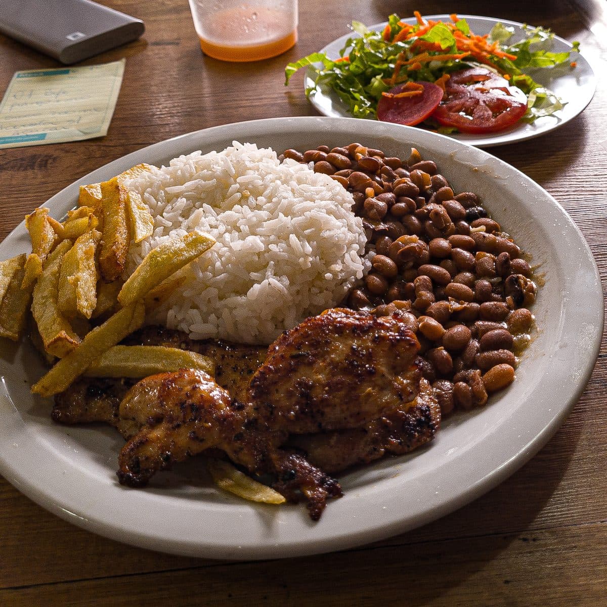 Prato feito com arroz, feijão, frango grelhado e batata frita, com salada ao fundo